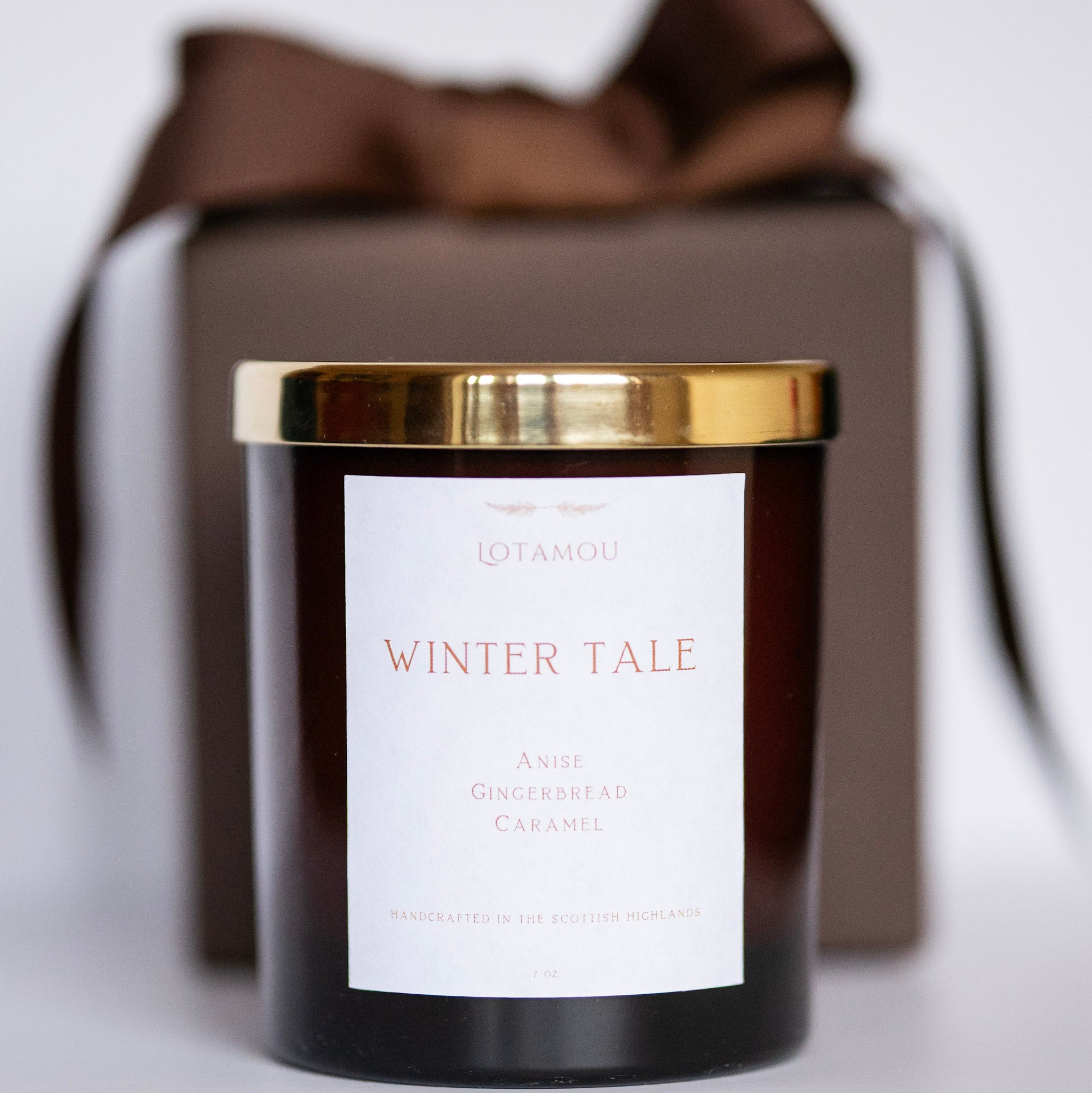 Candle with a gold lid and 'Winter Tale' label in front of a brown gift box.