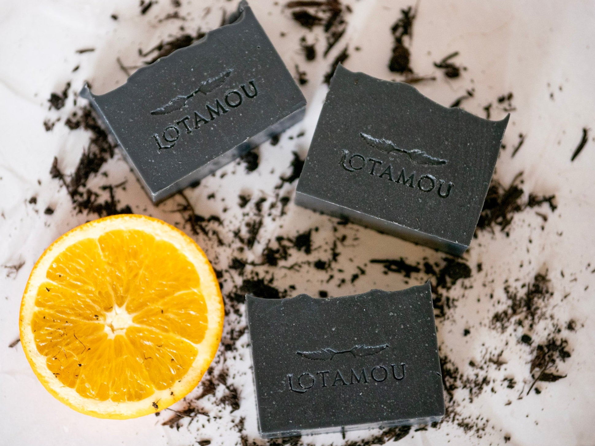 Three black soap bars with 'LOTAMOU' branding on a white surface with an orange slice.