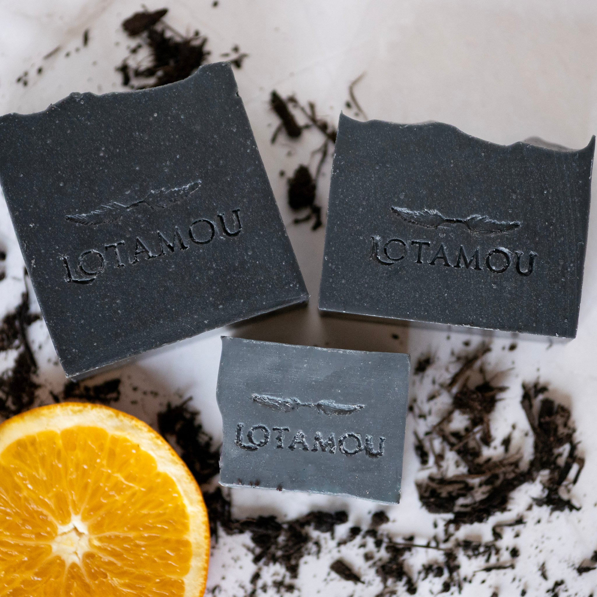 Three black soap bars with 'LOTAMOU' branding on a white surface with an orange half.
