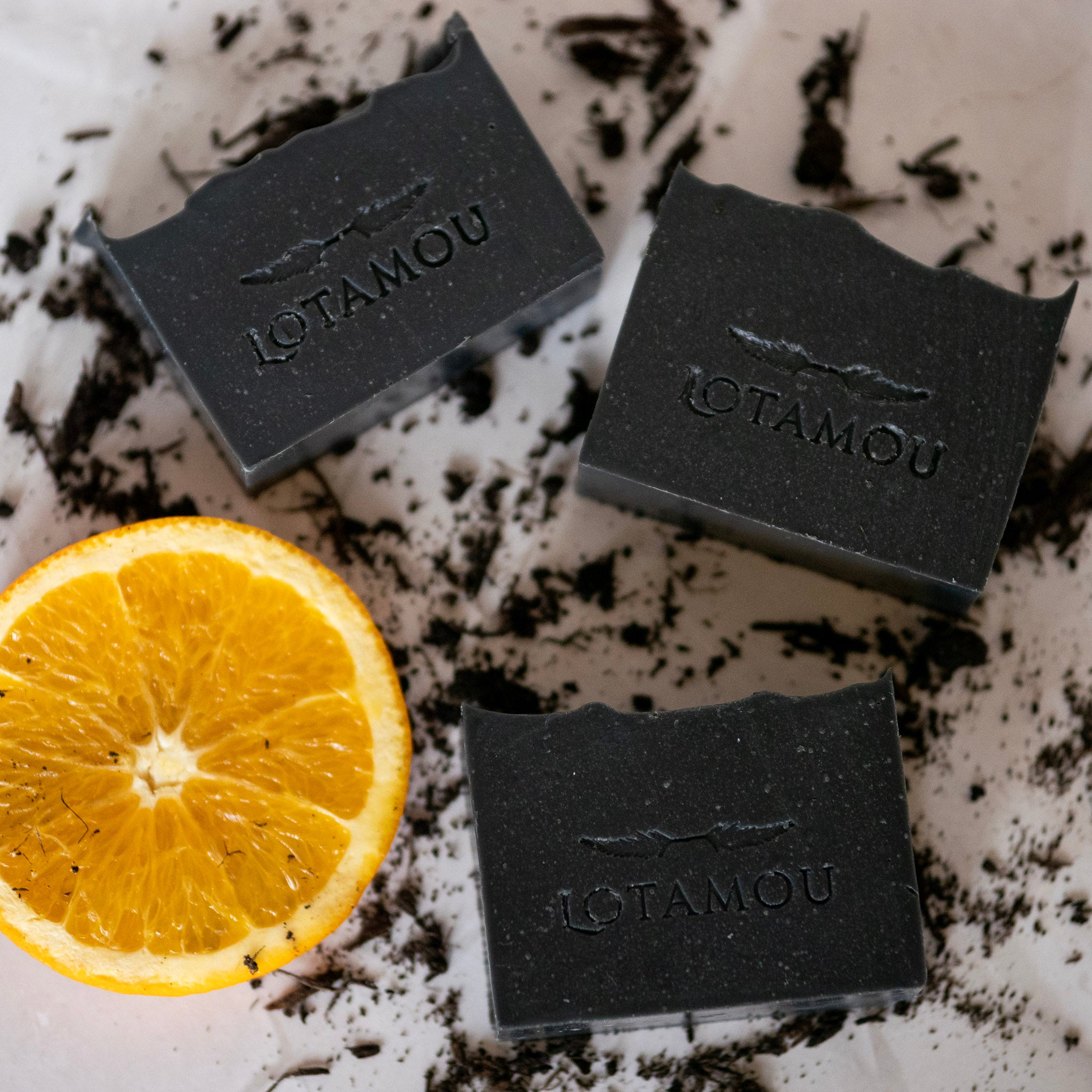 Three black soap bars with 'LOTAMOU' branding next to a lemon on a textured surface.