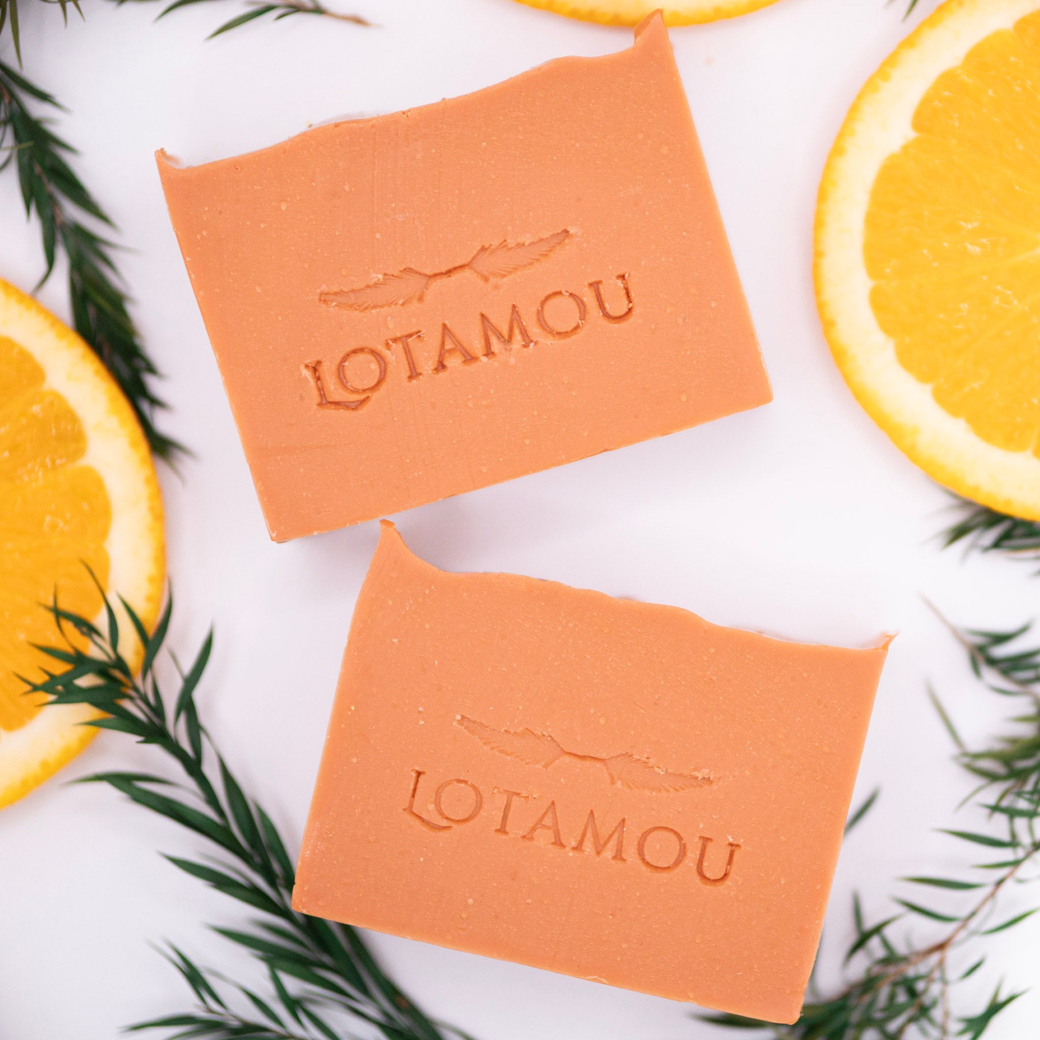Two orange soap bars with 'LOTAMOU' branding surrounded by orange slices and green leaves on a white background.