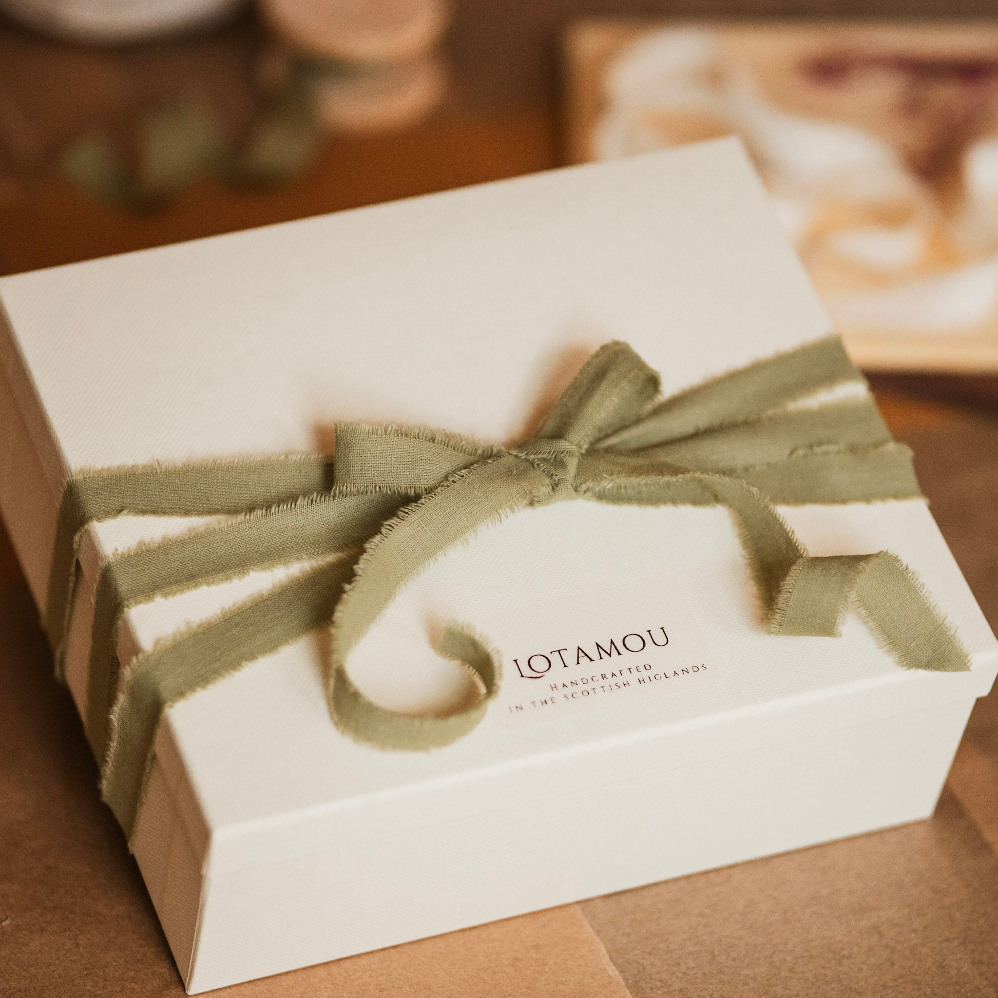 White gift box with a green ribbon and 'LOTAMOU' branding on a wooden surface.